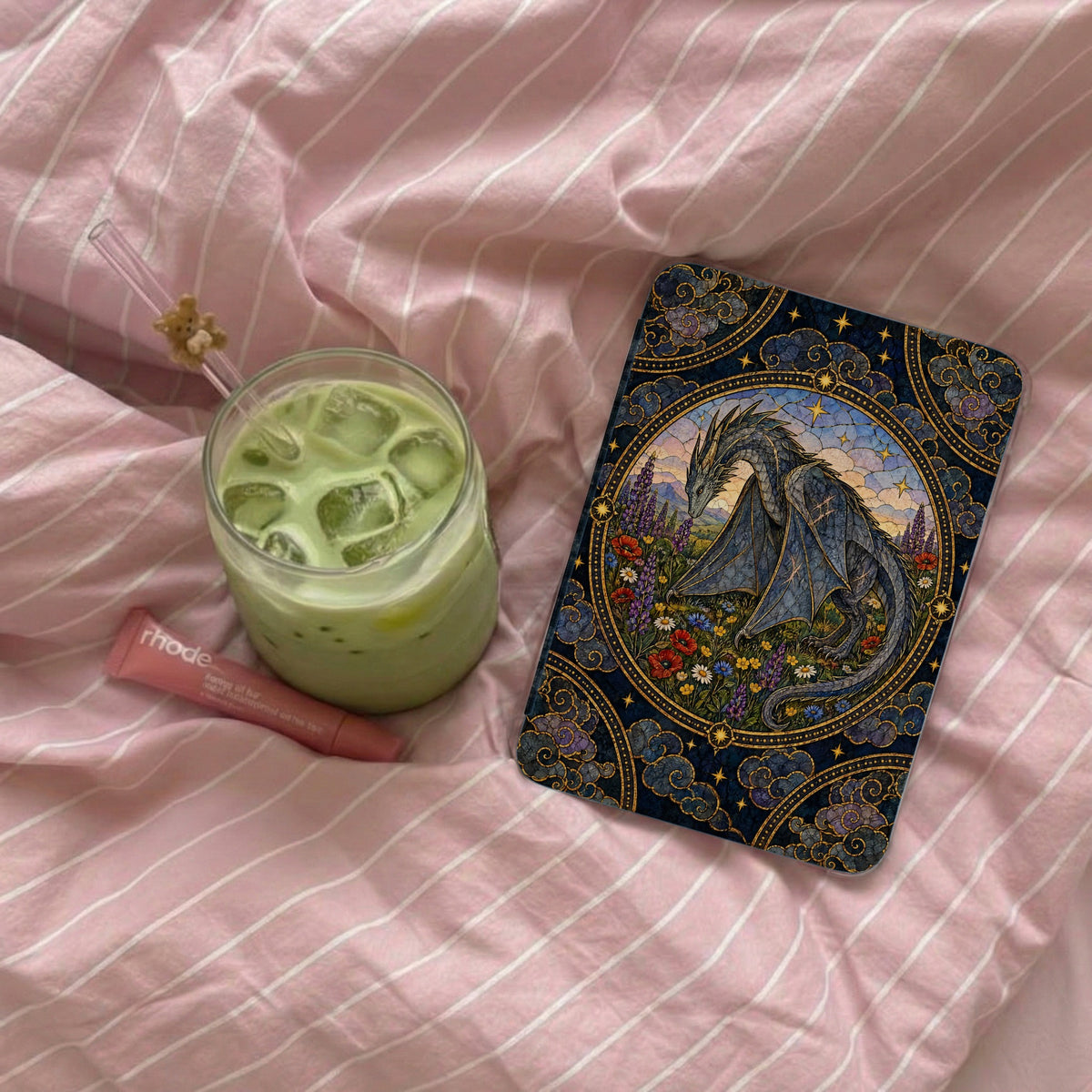 An e-reader with an ornate dragon case, an iced matcha latte, and a pink lip balm rest on a pink striped bedsheet.