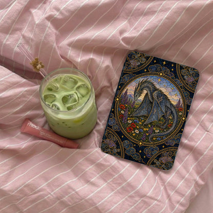 An e-reader with an ornate dragon case, an iced matcha latte, and a pink lip balm rest on a pink striped bedsheet.