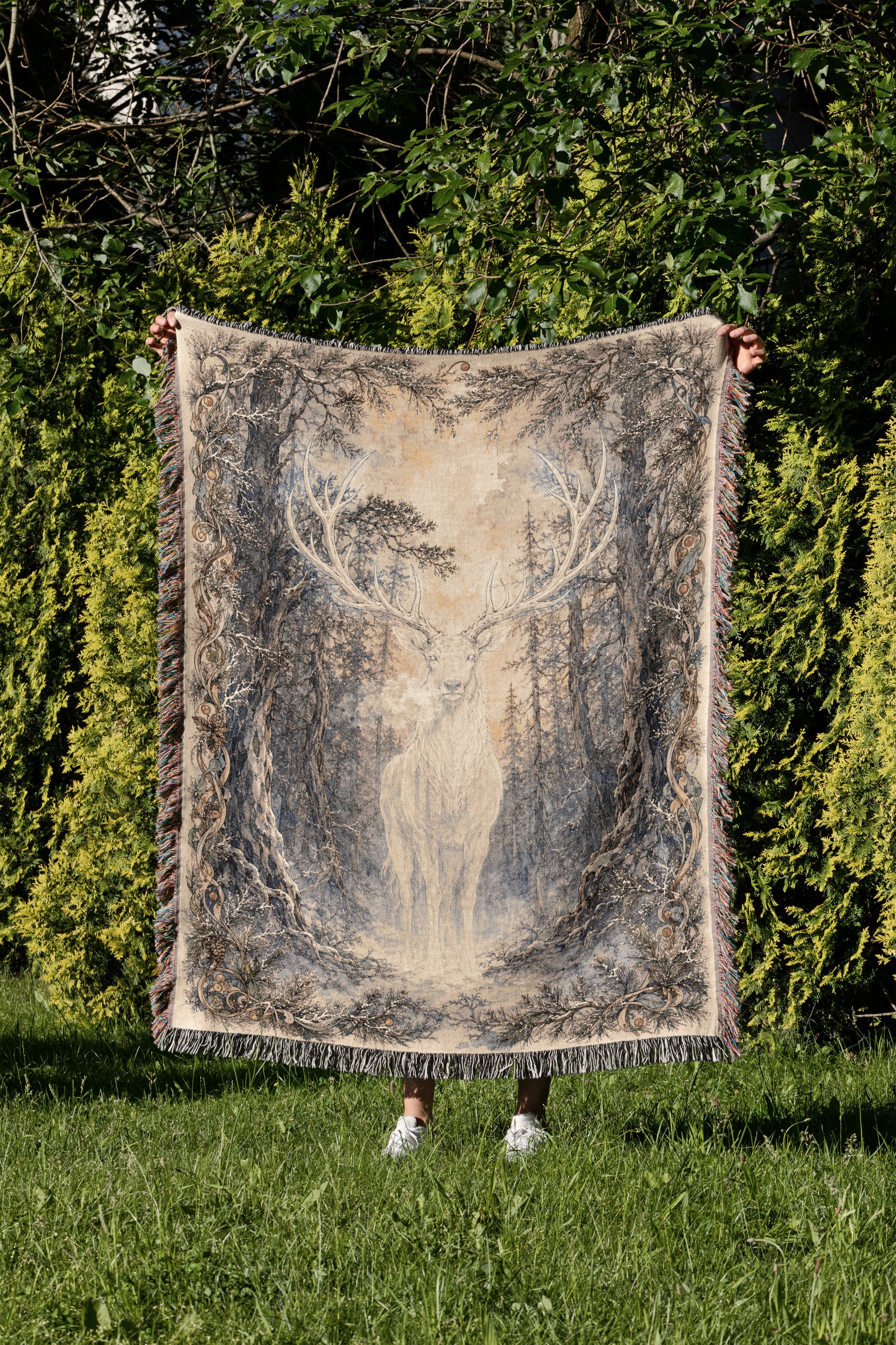A person is holding up a large blanket with a deer design on it, standing in a grassy area surrounded by trees.