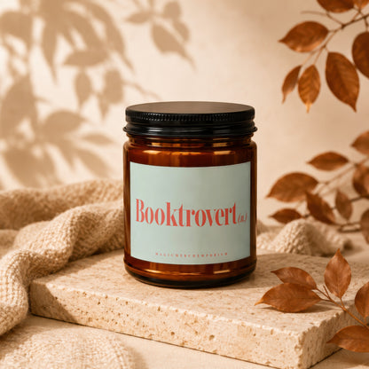 Amber jar candle with "Booktrovert" label on a stone block with fabric and leaves.
