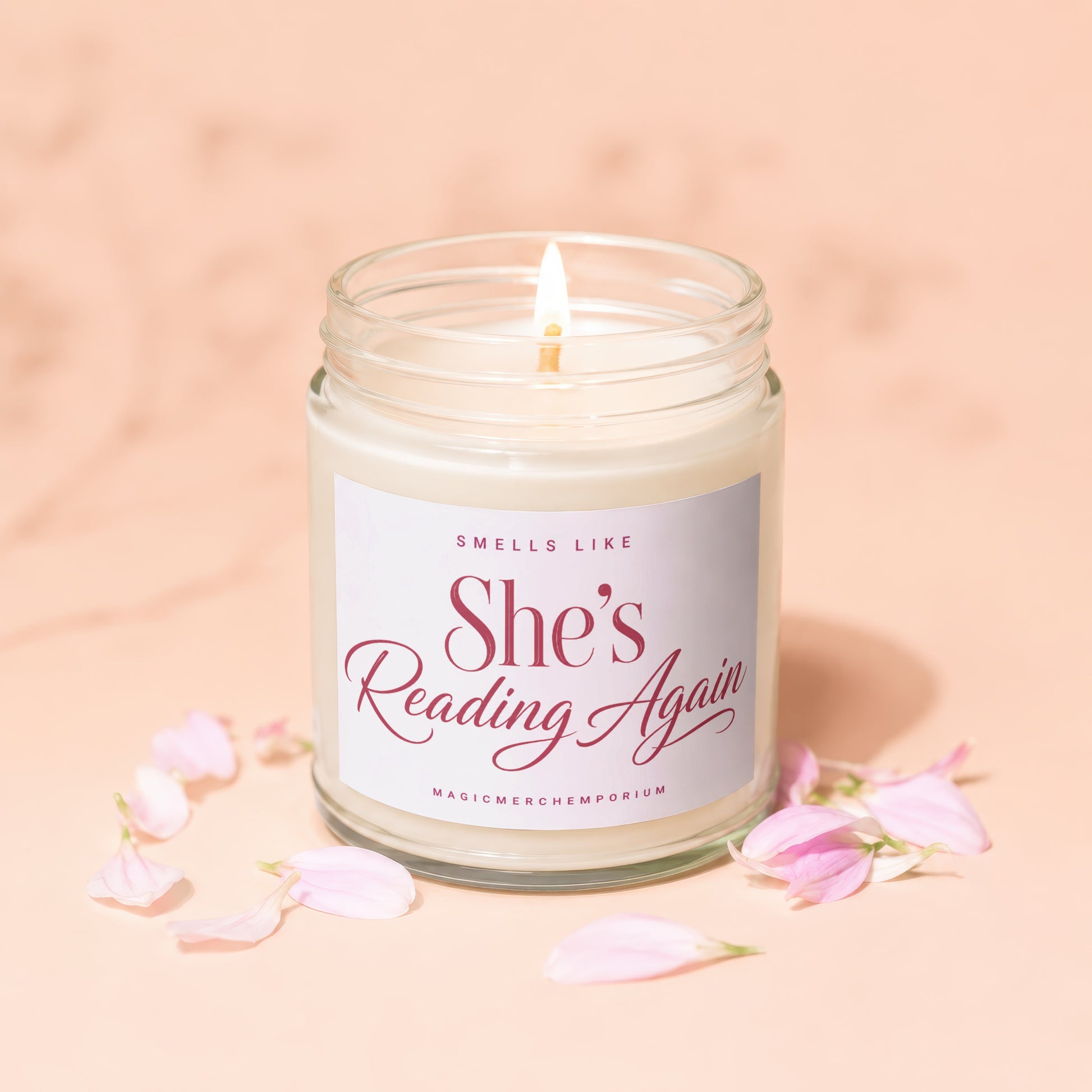 Candle with a lit wick on a pink marble surface surrounded by petals, labeled "She's Reading Again."