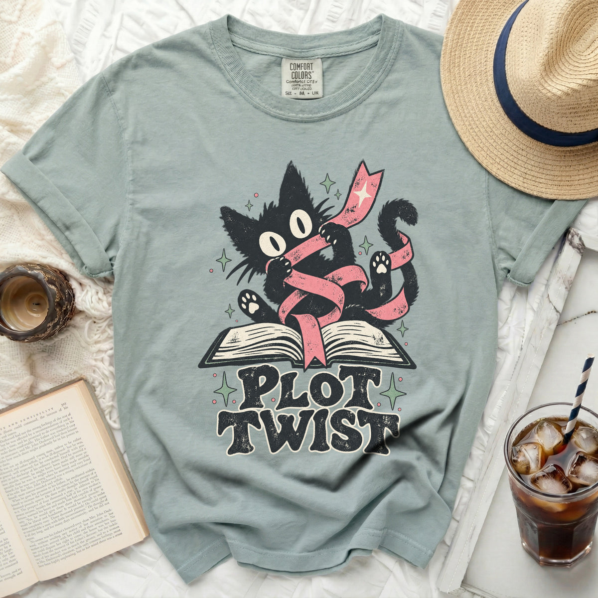 Graphic t-shirt featuring a black cat with large eyes wrapped in a pink ribbon over an open book, with text "PLOT TWIST".