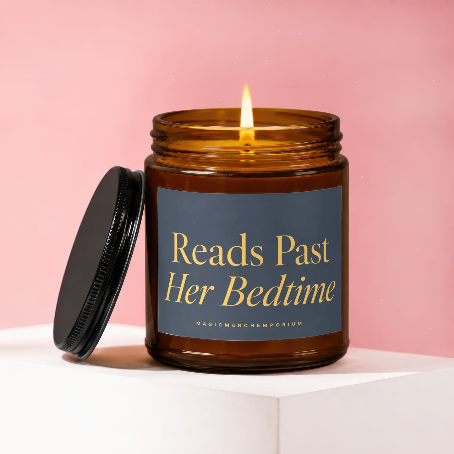 Lit amber glass candle with a black lid on a white surface against a pink background.