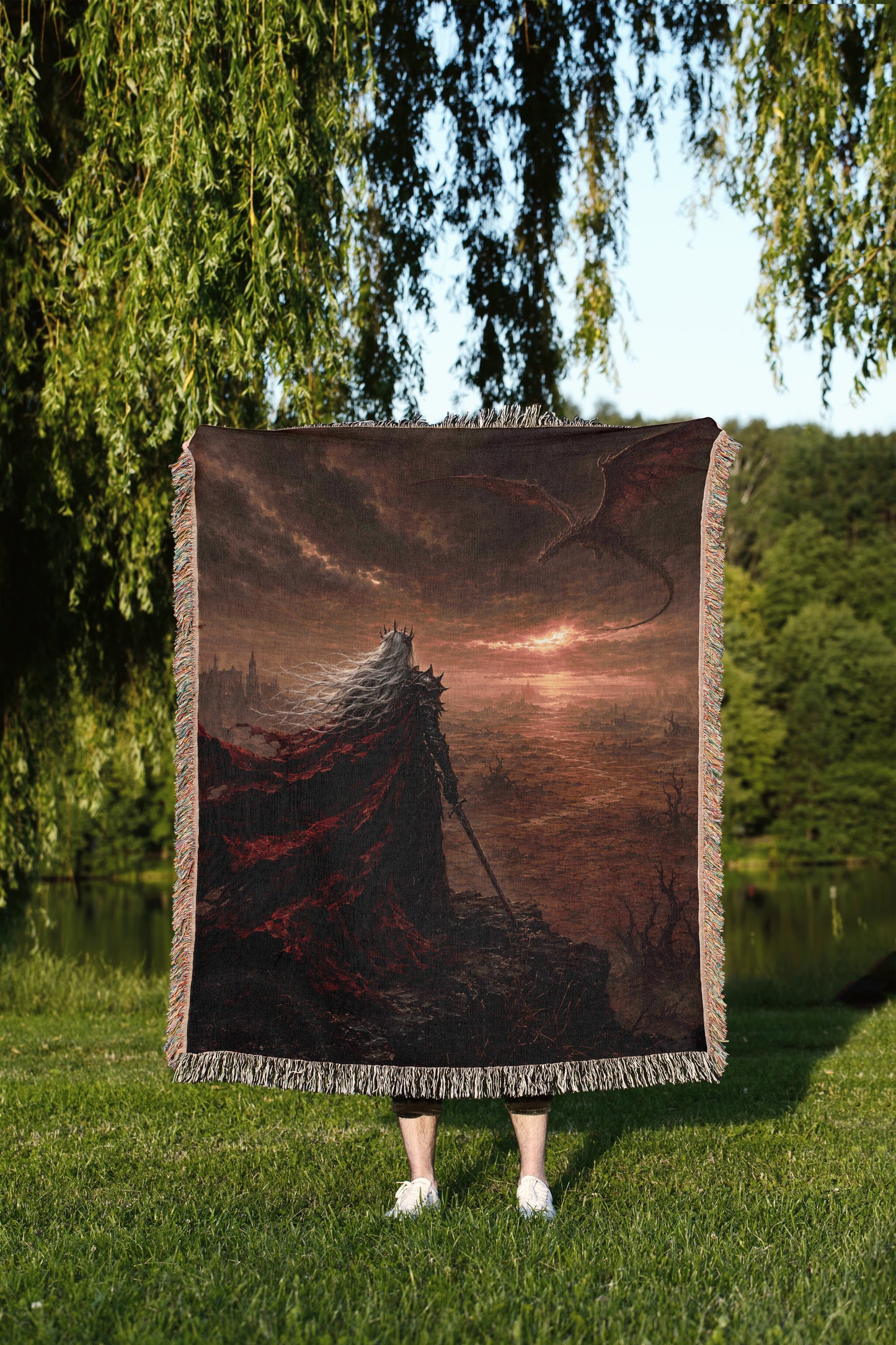 A person is standing in a grassy area, holding a large square blanket that features a painting of a dragon and a castle.
