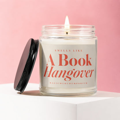 Glass candle with lit wick and black lid, reading "Smells Like A Book Hangover".