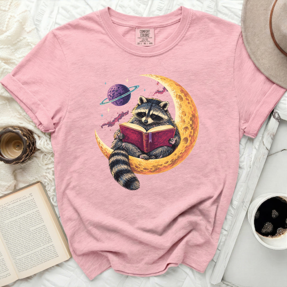 Pink t-shirt with graphic of raccoon reading book on crescent moon with planet.