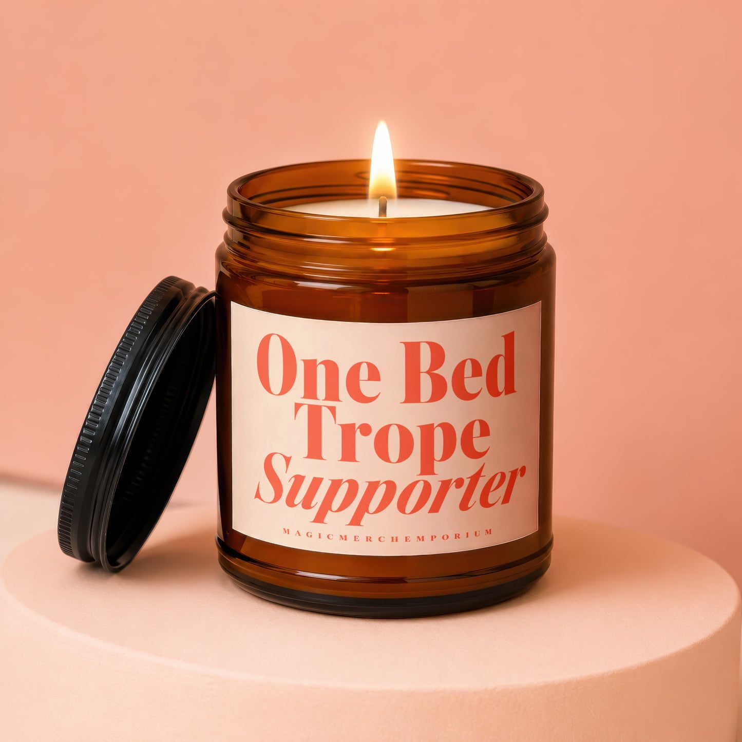 Amber jar candle with lit flame and black lid, reads "One Bed Trope Supporter".