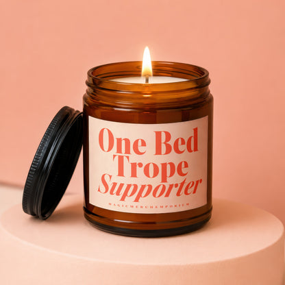 Amber jar candle with lit flame and black lid, reads "One Bed Trope Supporter".