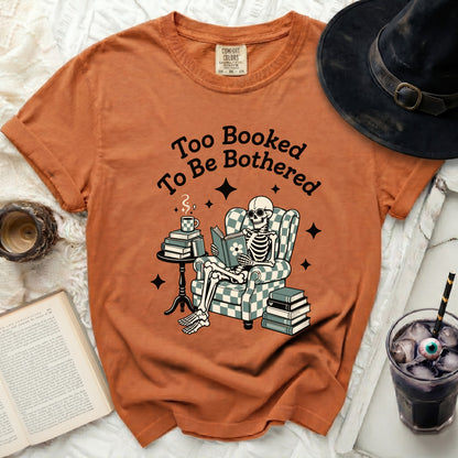 Orange t-shirt with a skeleton reading a book and text "Too Booked To Be Bothered."