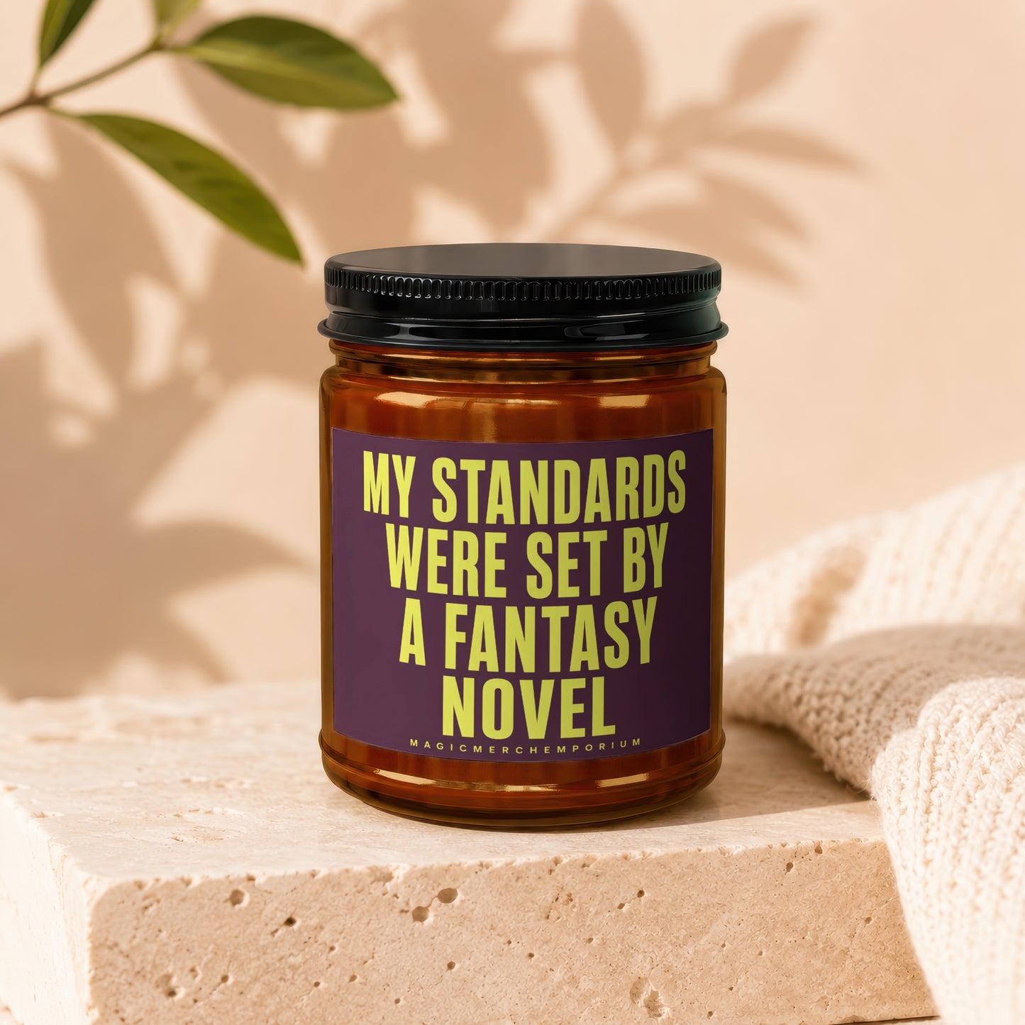Brown glass candle with black lid features a purple label with yellow text reading "MY STANDARDS WERE SET BY A FANTASY NOVEL".
