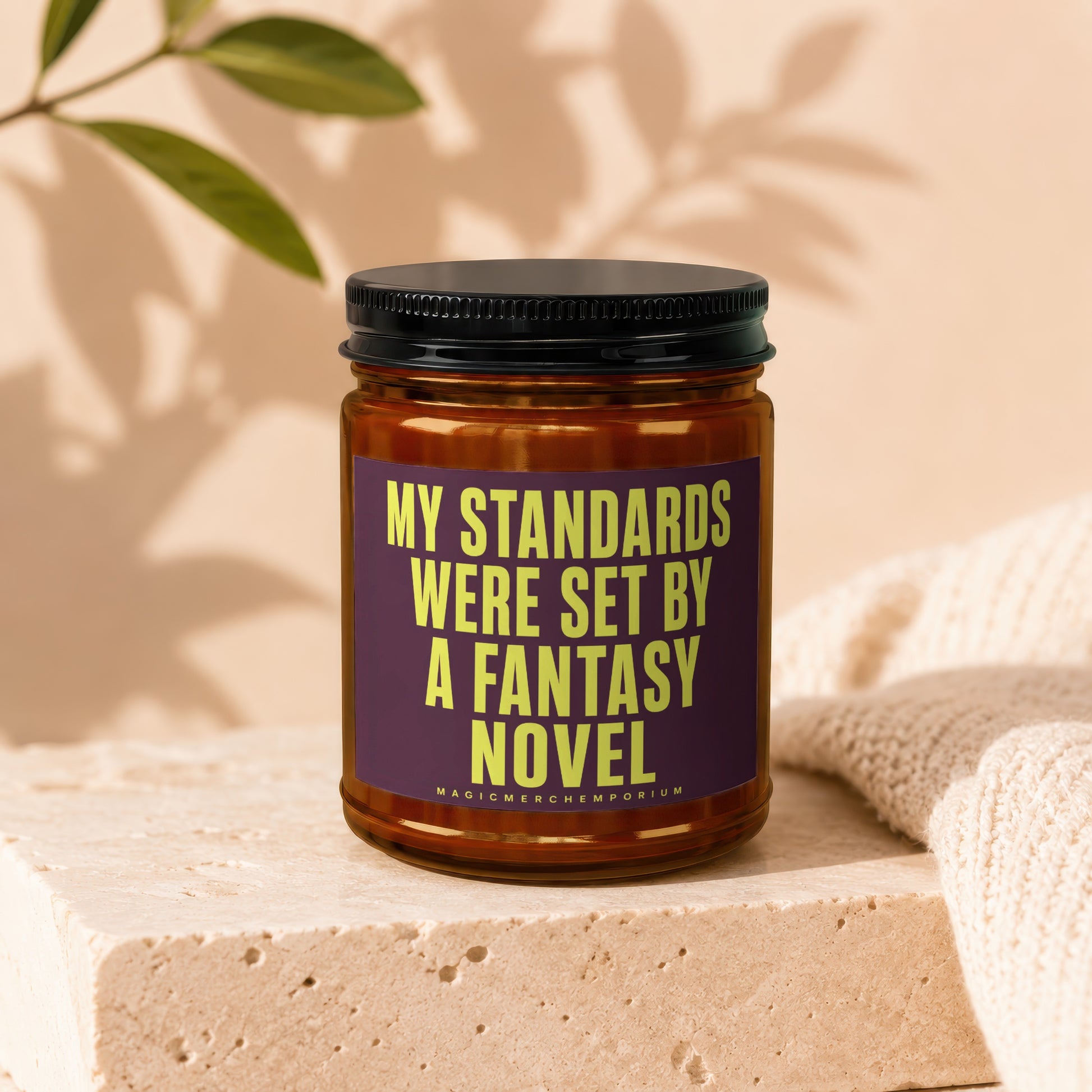 Brown glass candle with black lid features a purple label with yellow text reading "MY STANDARDS WERE SET BY A FANTASY NOVEL".