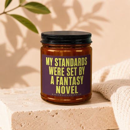 Brown glass candle with black lid features a purple label with yellow text reading "MY STANDARDS WERE SET BY A FANTASY NOVEL".