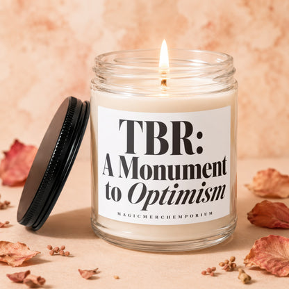 Candle with lid off, displaying text "TBR: A Monument to Optimism" and brand.