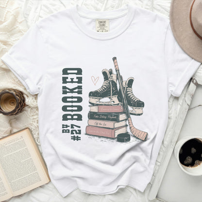 White t-shirt with stacked books, ice skates, and hockey stick graphic.