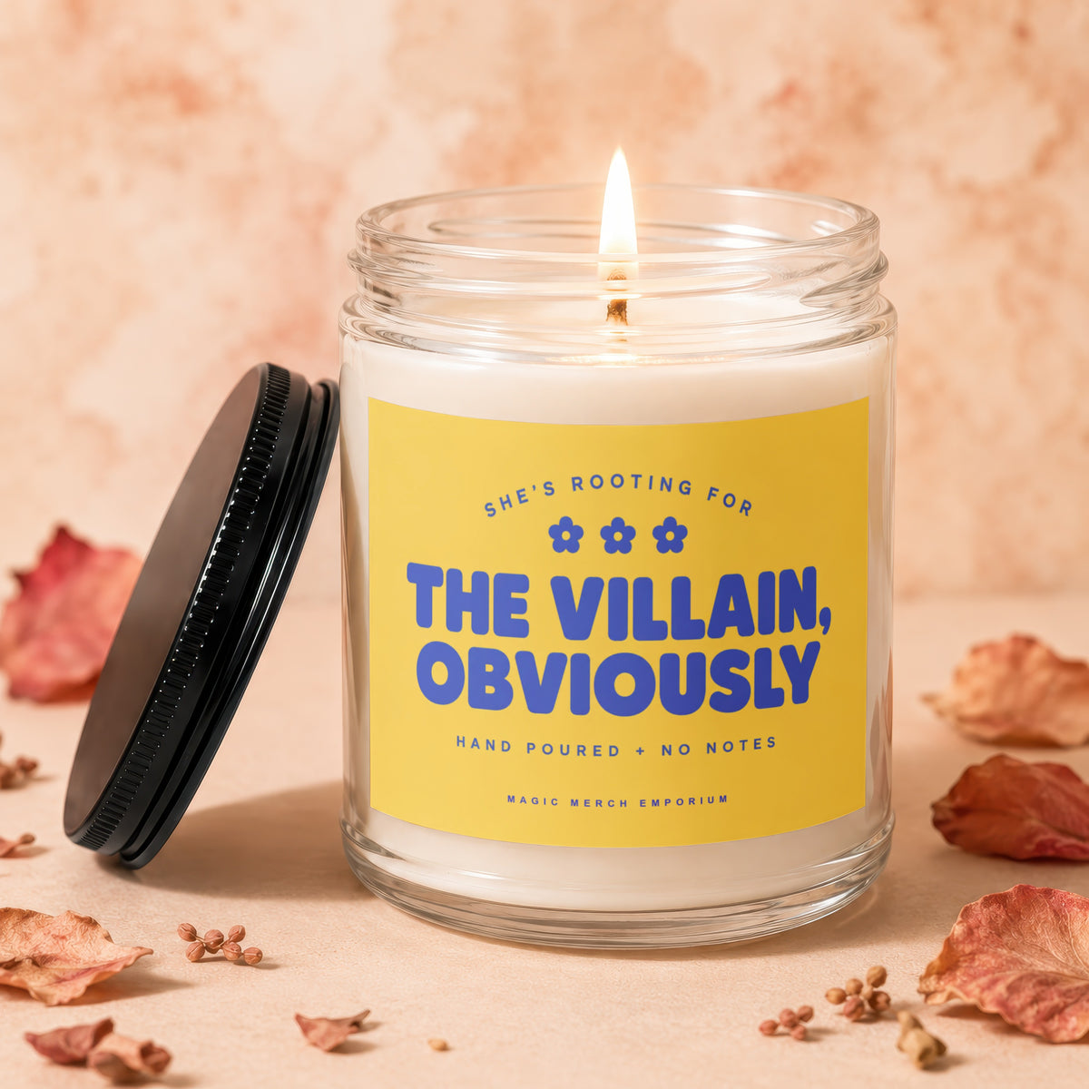 Lit candle in glass jar with yellow label reading "SHE'S ROOTING FOR THE VILLAIN, OBVIOUSLY."