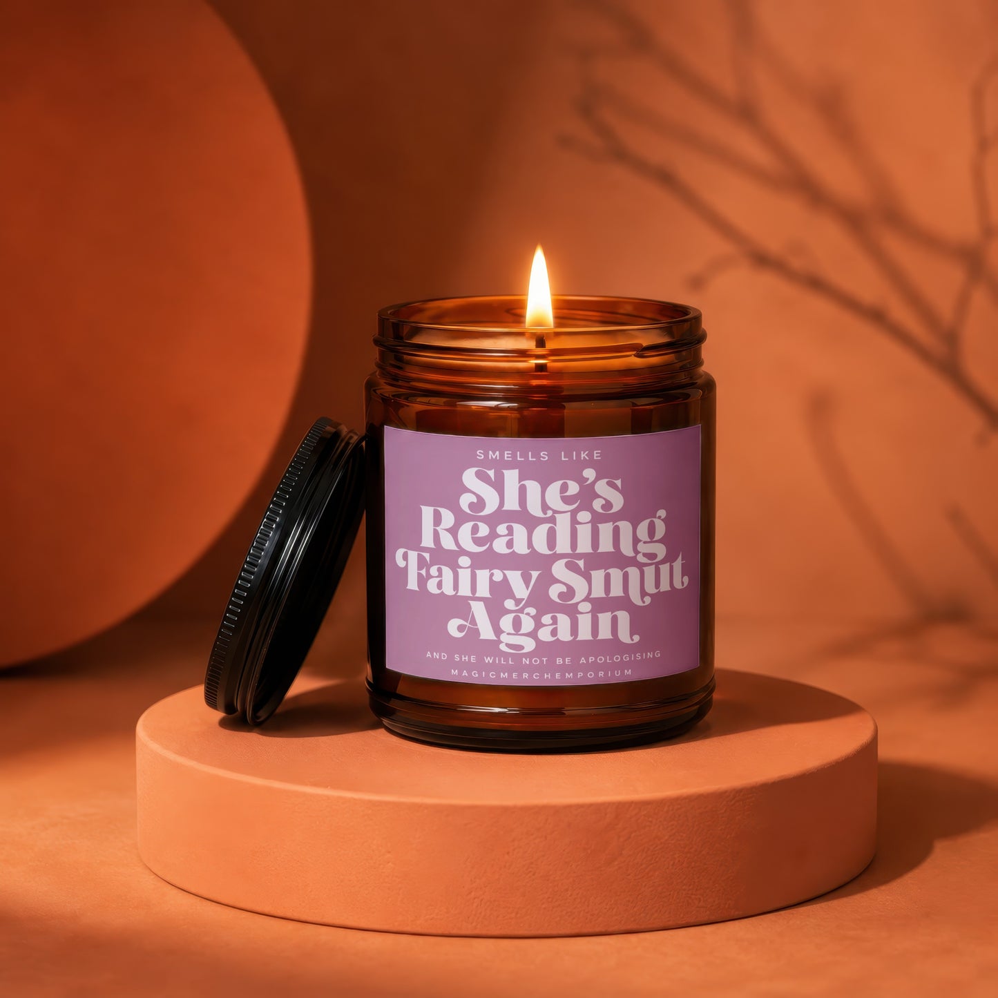 Amber jar candle with lit flame and purple label reading "She's Reading Fairy Smut Again".