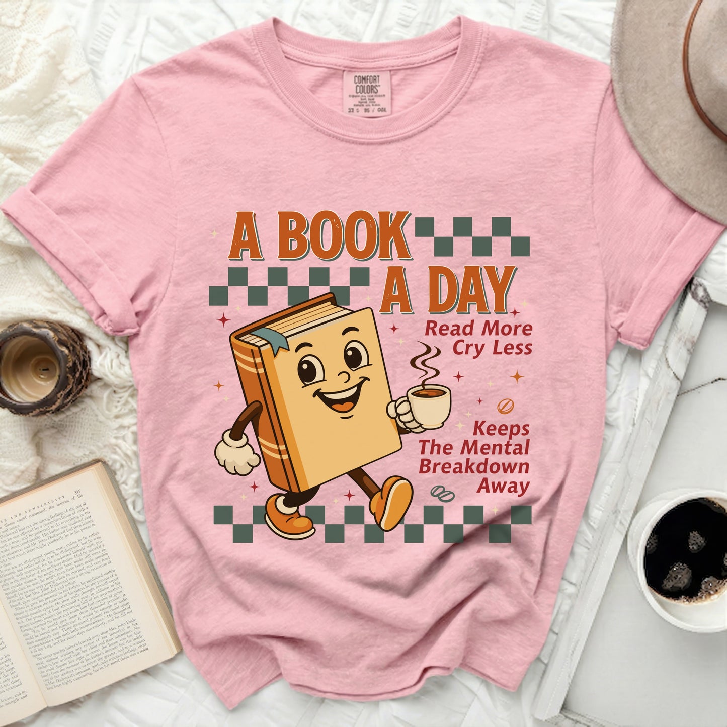 Pink t-shirt with a cartoon book holding coffee, reading "A book a day, Read more, Cry less, Keeps the mental breakdown away."