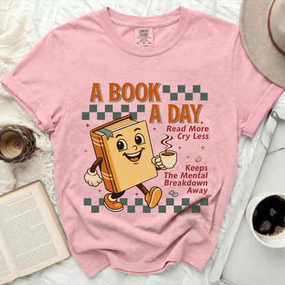Pink t-shirt with a cartoon book holding coffee, reading "A book a day, Read more, Cry less, Keeps the mental breakdown away."