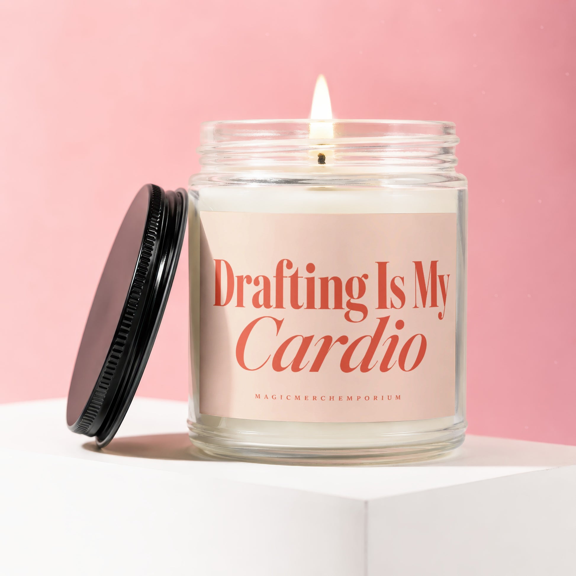 Lit jar candle with pink label reading "Drafting Is My Cardio" on pink background.