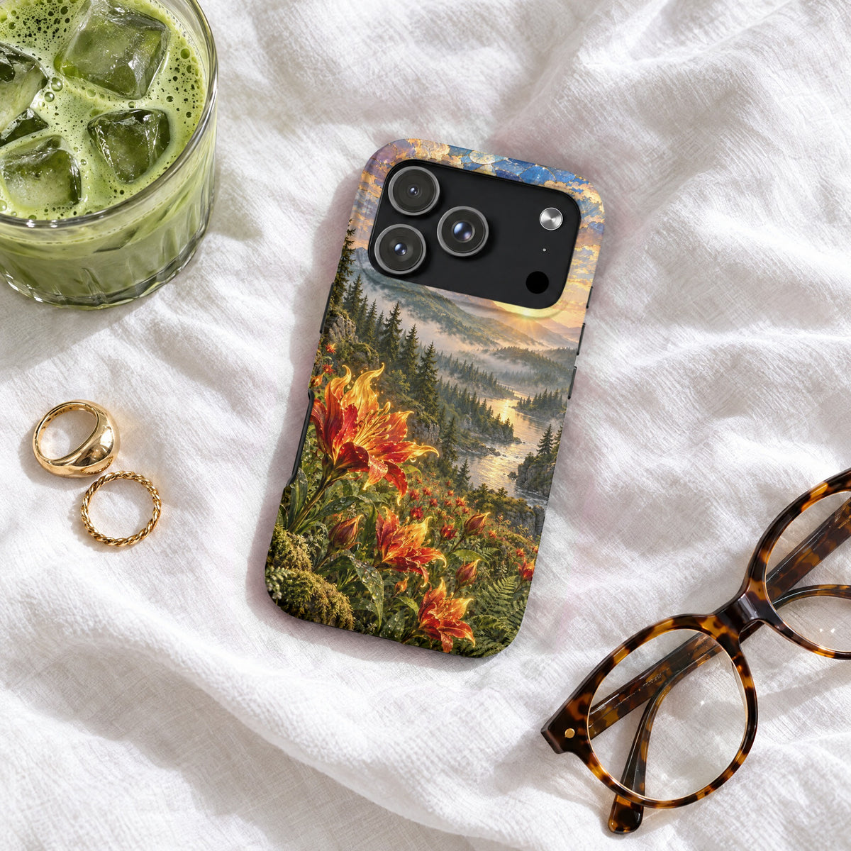 A smartphone case featuring a landscape print of a river valley with orange lilies on a white fabric surface with a glass of iced green tea, two gold rings, and tortoiseshell glasses.