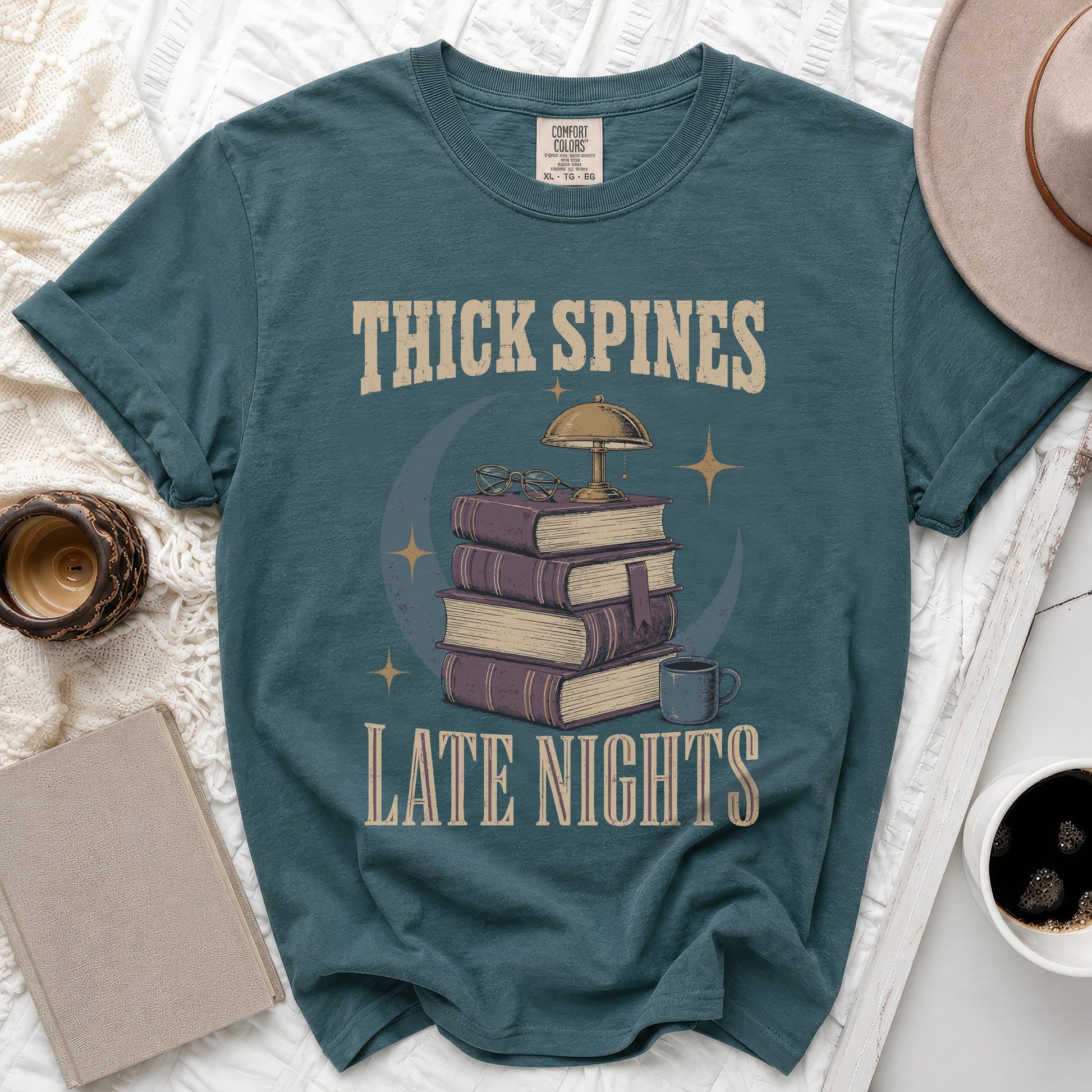 Teal t-shirt with "THICK SPINES LATE NIGHTS" text, books, lamp, and mug graphic.
