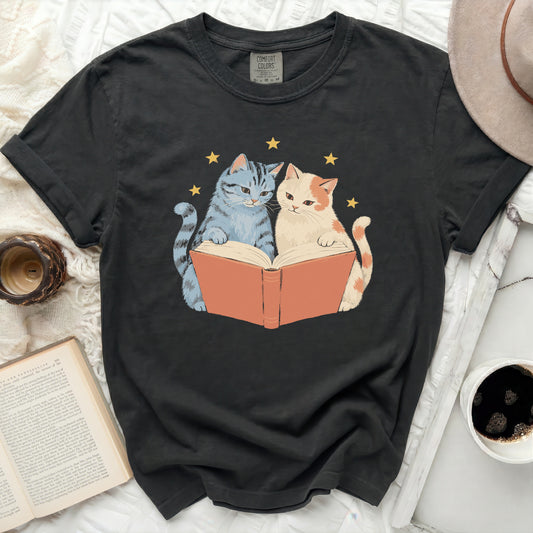 T-shirt with a graphic of two cats reading a book under stars.