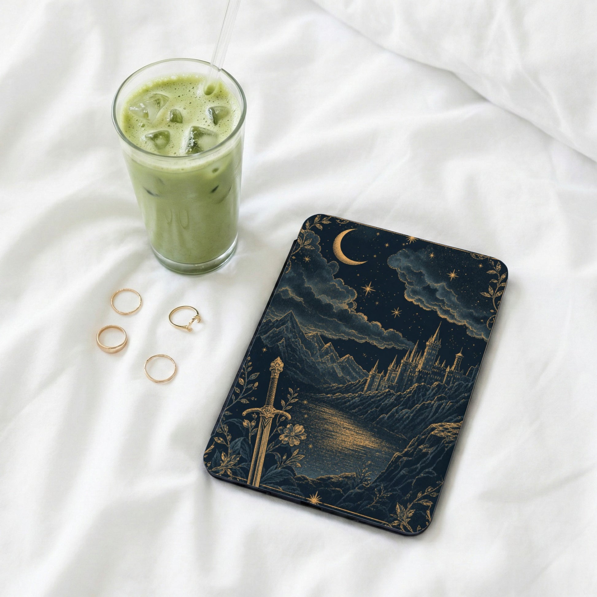 Book with fantasy castle art, sword, moon, and stars, next to iced matcha drink and rings.