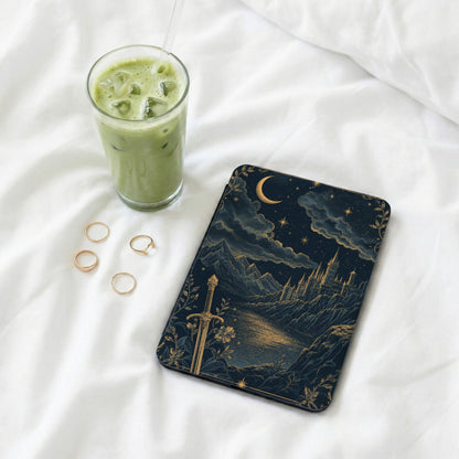 Book with fantasy castle art, sword, moon, and stars, next to iced matcha drink and rings.