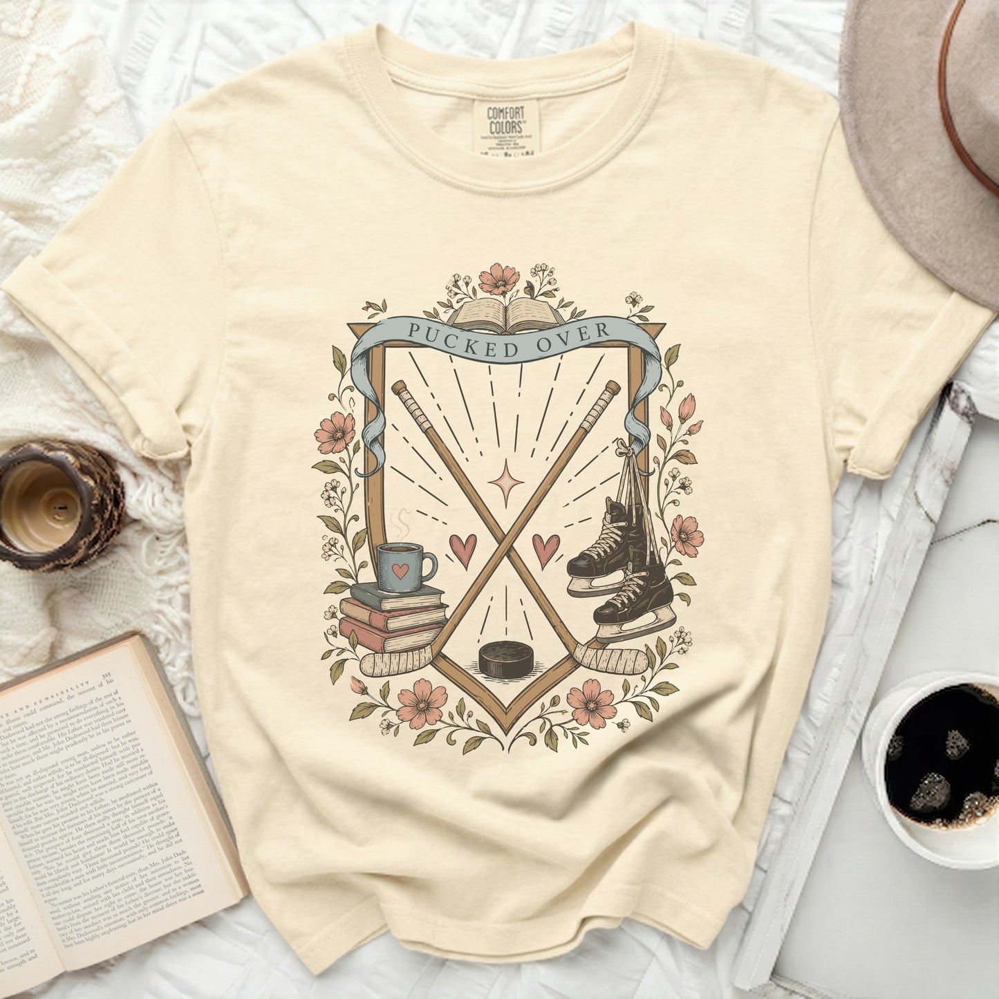 Cream t-shirt with "Pucked Over" crest, hockey sticks, skates, and books.