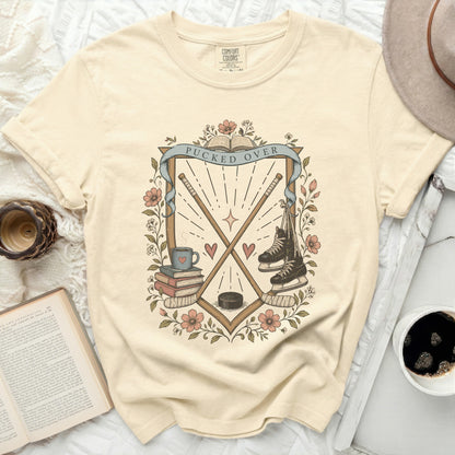 Cream t-shirt with "Pucked Over" crest, hockey sticks, skates, and books.
