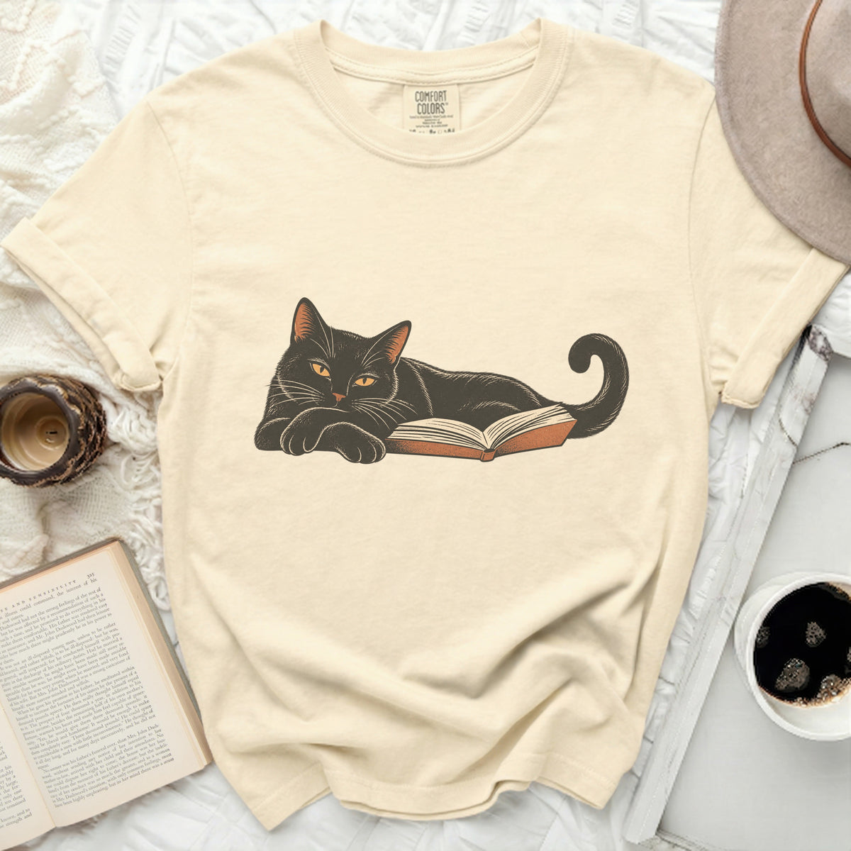 Black cat lying on an open book, Comfort Colors tag visible.