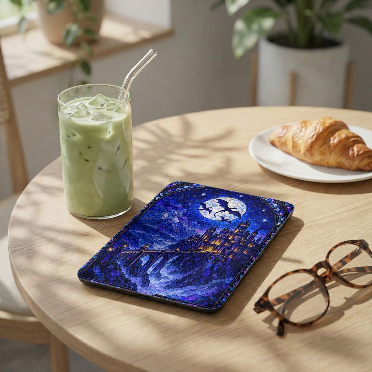 E-reader case with a blue stained-glass castle design rests on a wooden table beside a green iced drink, croissant, and glasses.