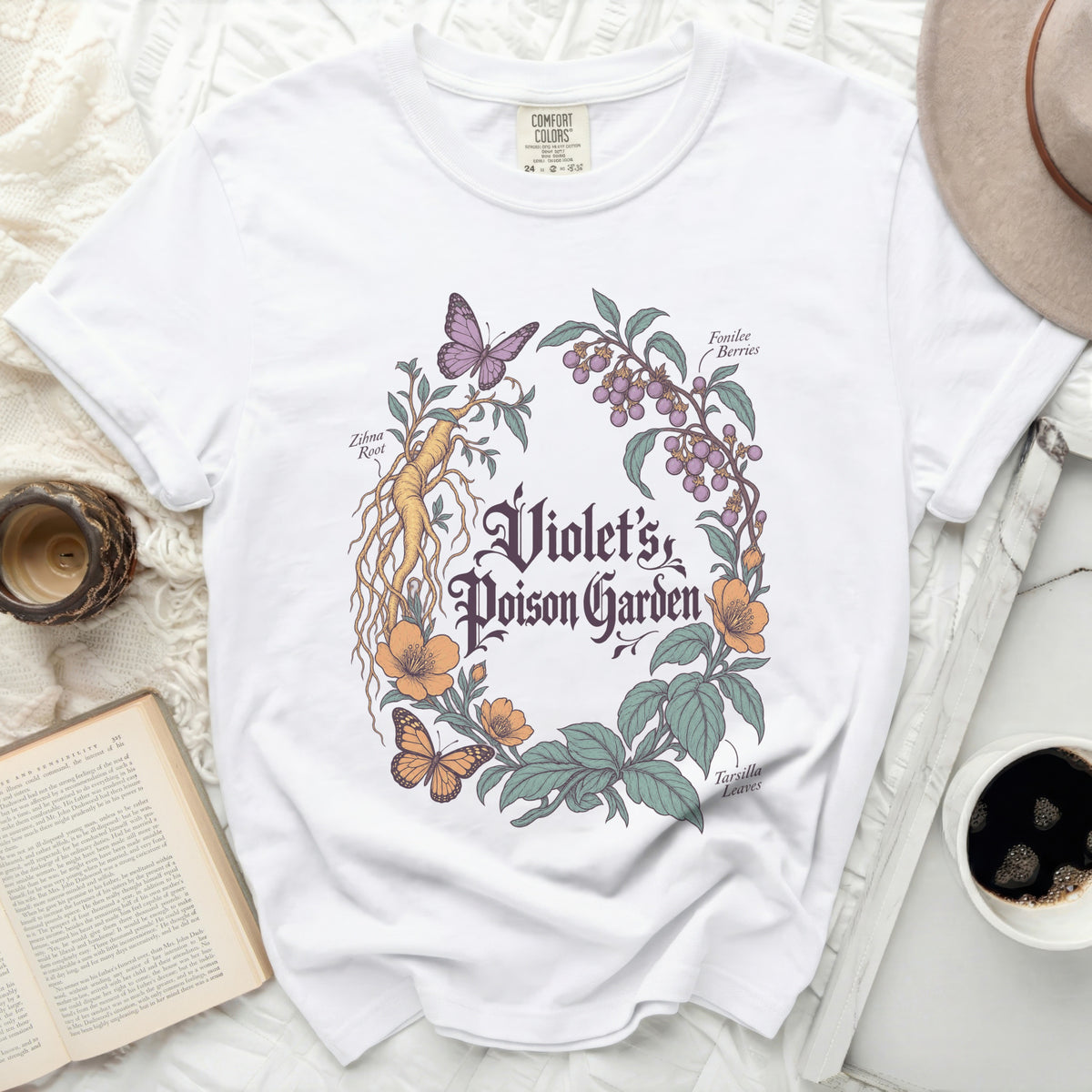 A white t-shirt featuring a botanical illustration with text reading "Violet's Poison Garden" surrounded by plants, berries, and butterflies.