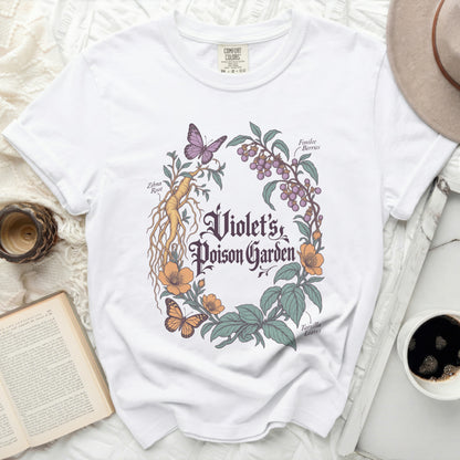 A white t-shirt featuring a botanical illustration with text reading "Violet's Poison Garden" surrounded by plants, berries, and butterflies.