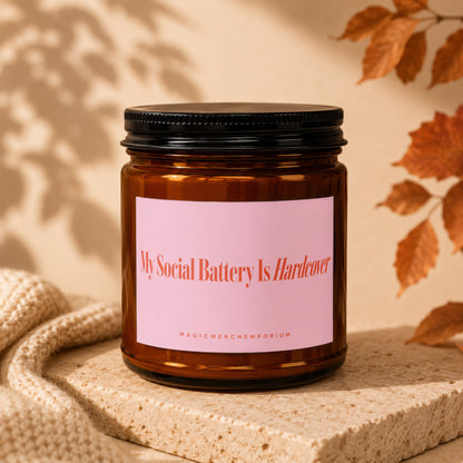 Amber candle in jar with pink label reading "My Social Battery Is Hardcover" and "MAGICMERCHEMPORIUM".