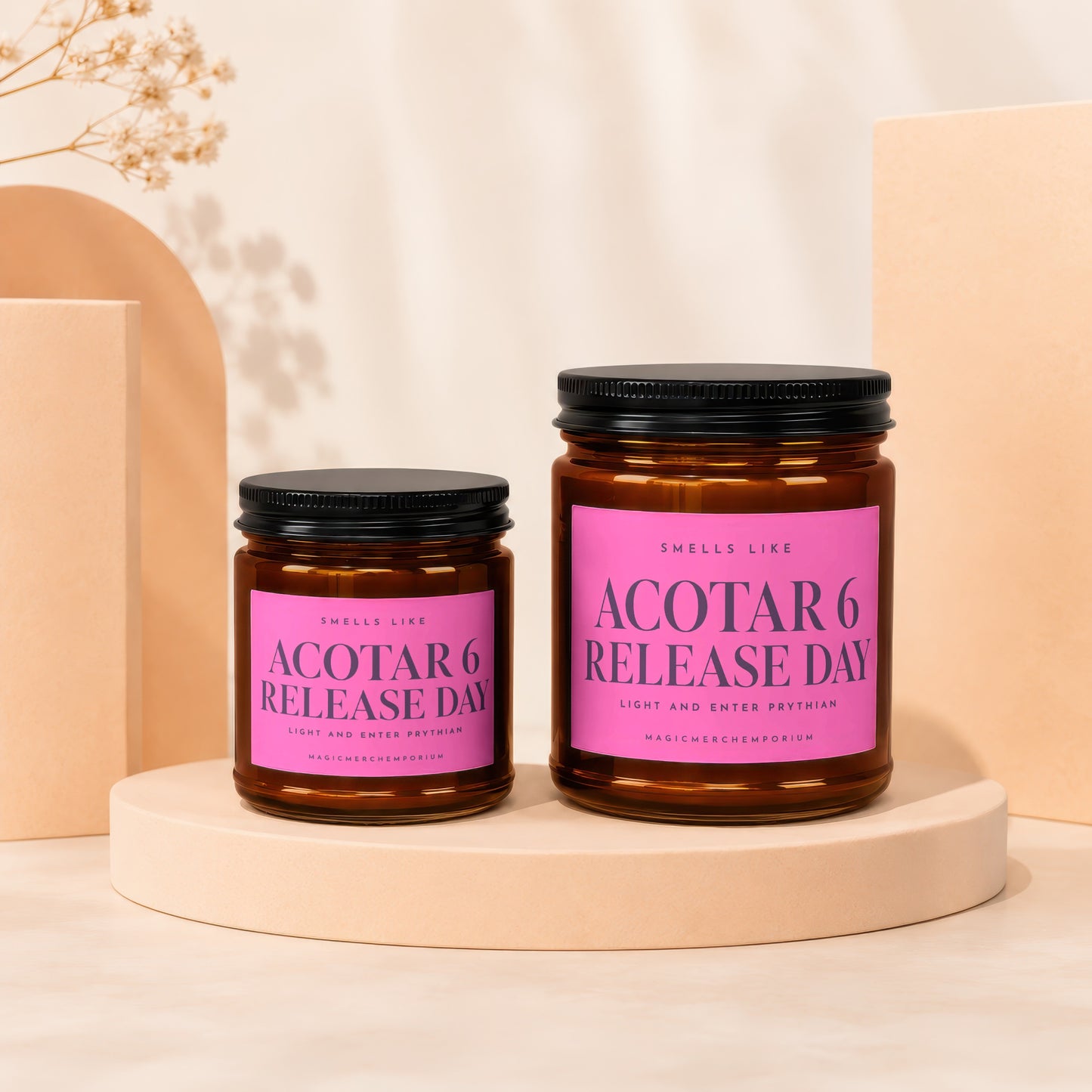 Two amber glass candle jars with pink labels reading "Smells Like ACOTAR 6 Release Day" on a beige platform.
