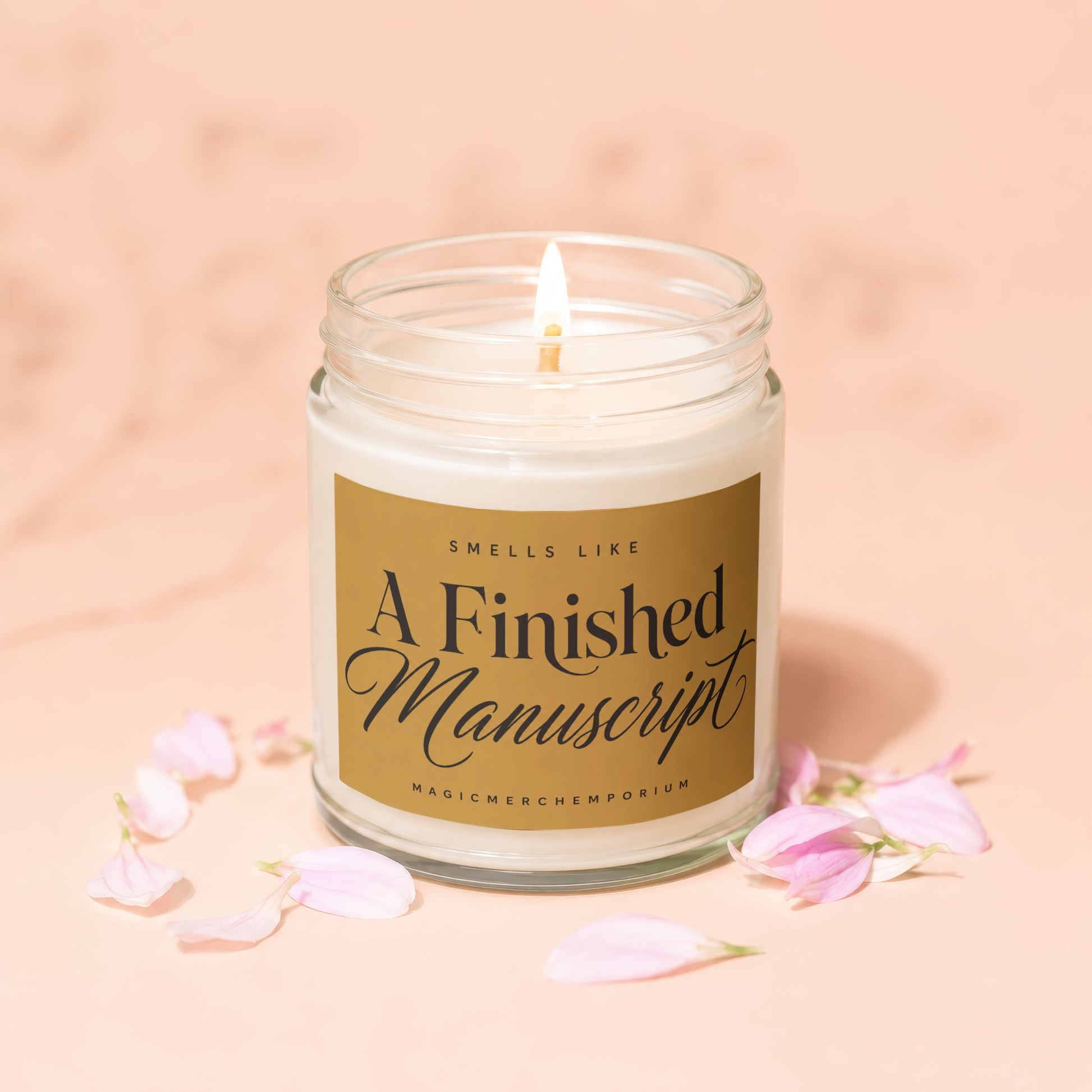 Lit candle in a glass jar with gold label reading "Smells Like A Finished Manuscript".