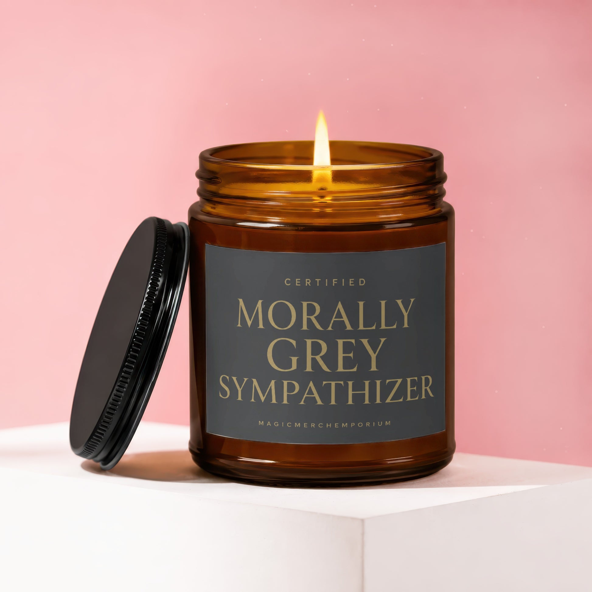 Amber jar candle labeled "Morally Grey Sympathizer" with lit flame on pink background.