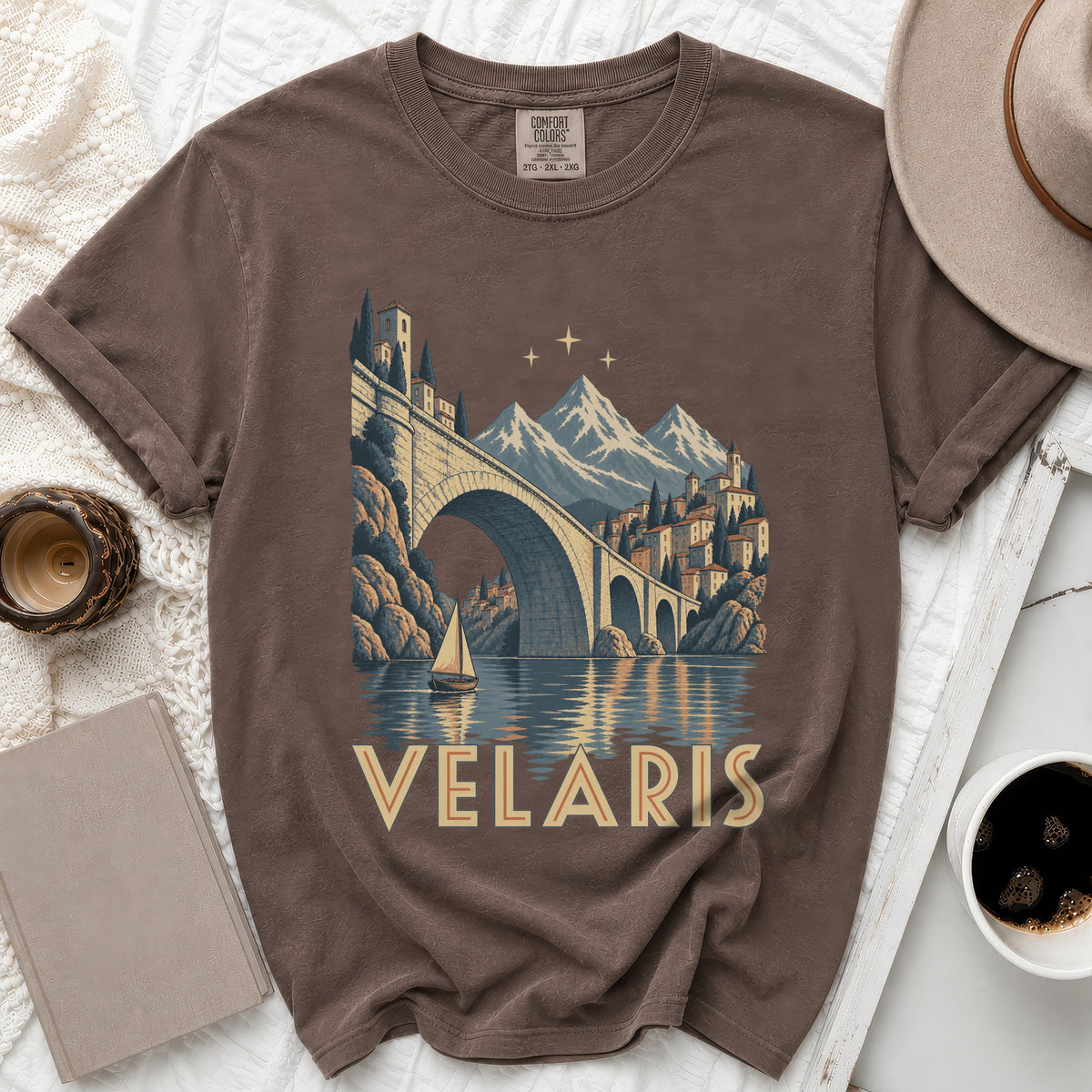 Brown t-shirt with graphic of Velaris cityscape, bridge, mountains, and sailboat.