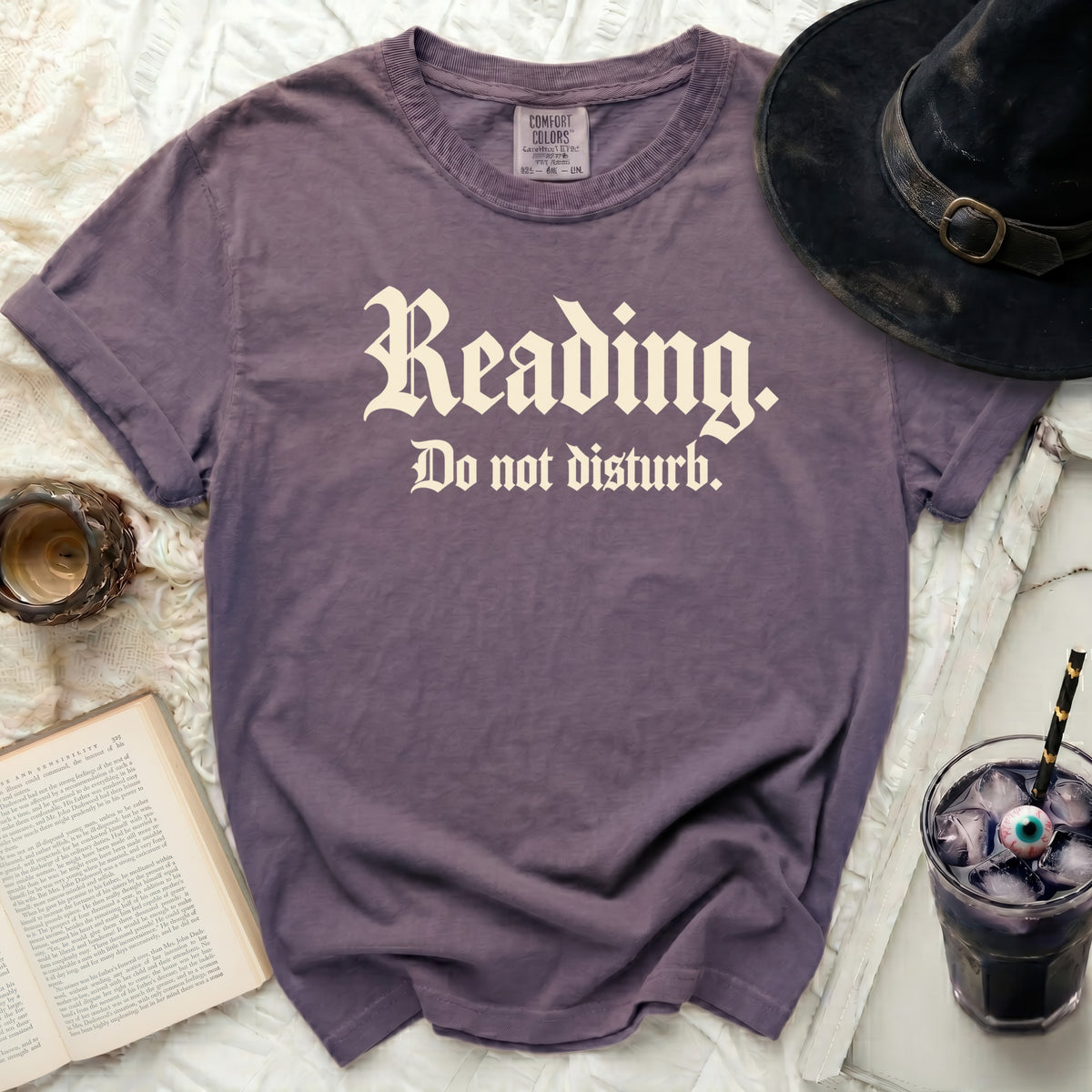 Purple t-shirt with "Reading. Do not disturb." text, witch hat, book, and drink.