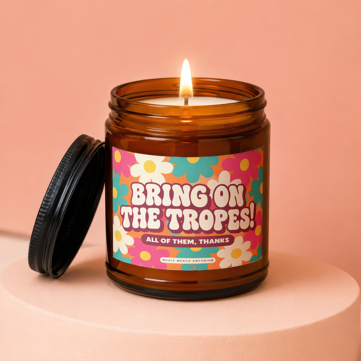 Amber glass candle with lid off, featuring a colorful floral label that reads "Bring on the Tropes! All of Them, Thanks".