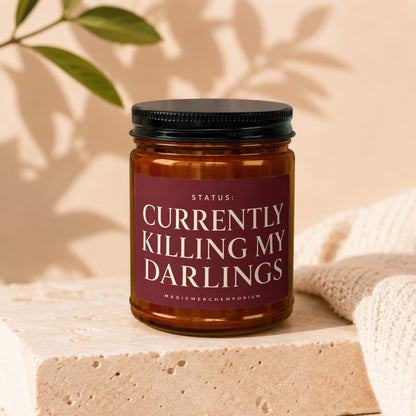 Amber jar candle with black lid reading "STATUS: CURRENTLY KILLING MY DARLINGS".