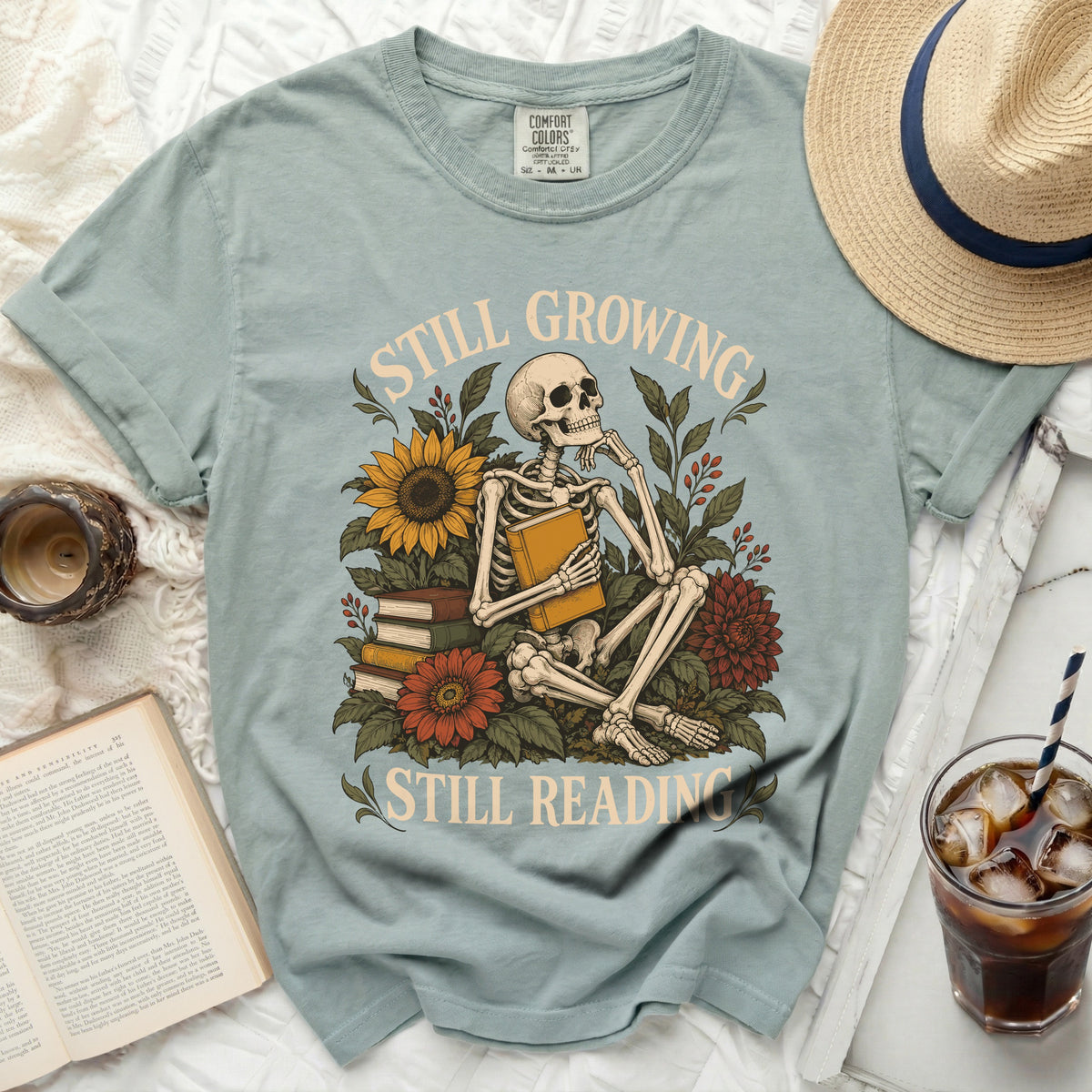 Skeleton reading a book surrounded by flowers with text "Still Growing Still Reading".