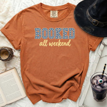 Orange t-shirt with "BOOKED all weekend" text, witch hat, book, and drink.
