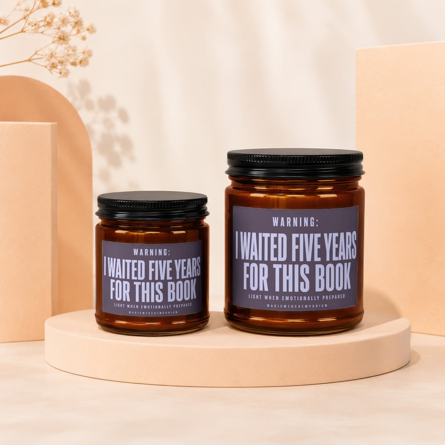 Two amber glass candle jars with black lids and labels reading I WAITED FIVE YEARS FOR THIS BOOK on a beige pedestal.