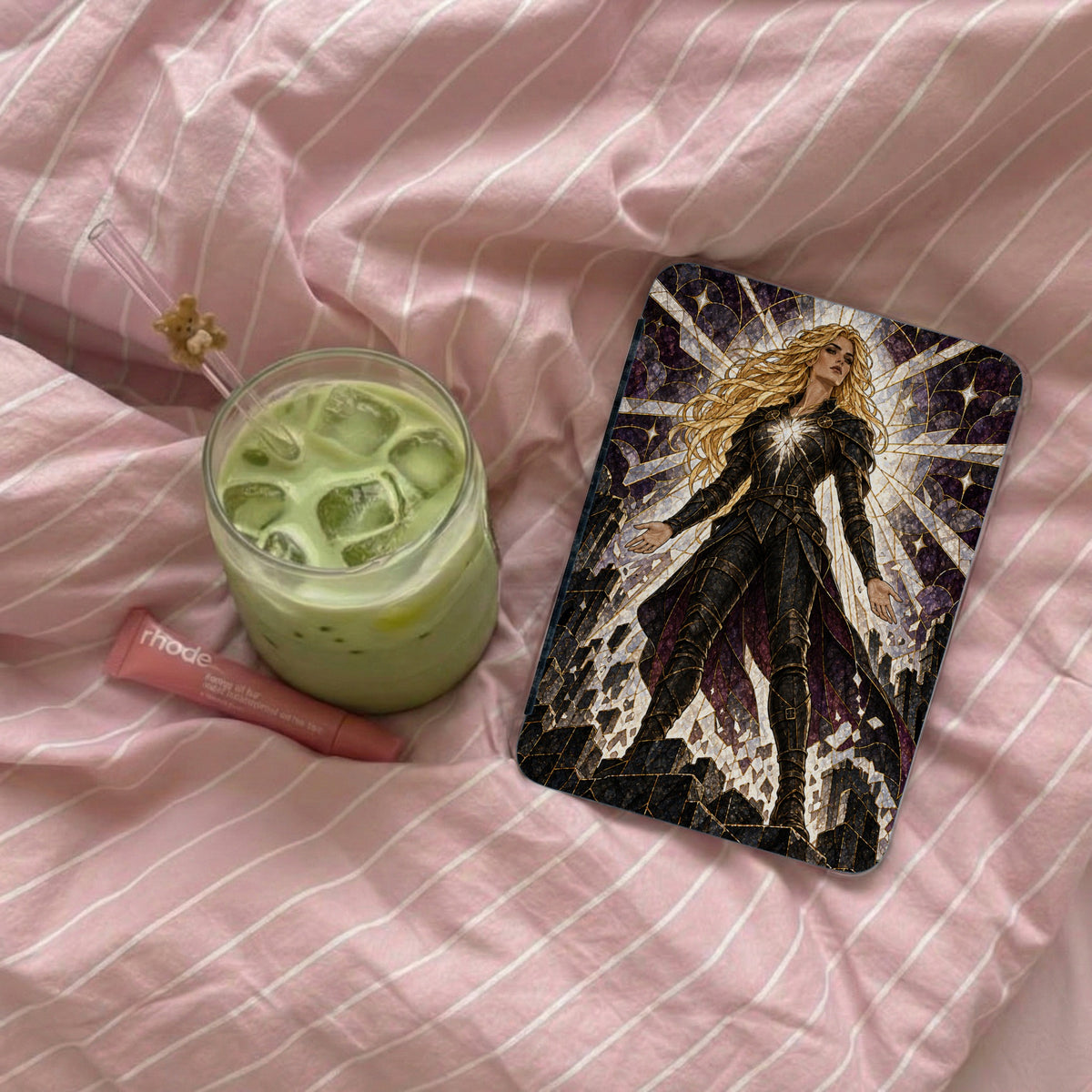 Case with blonde warrior artwork, glass of matcha, and lip balm on pink striped fabric.