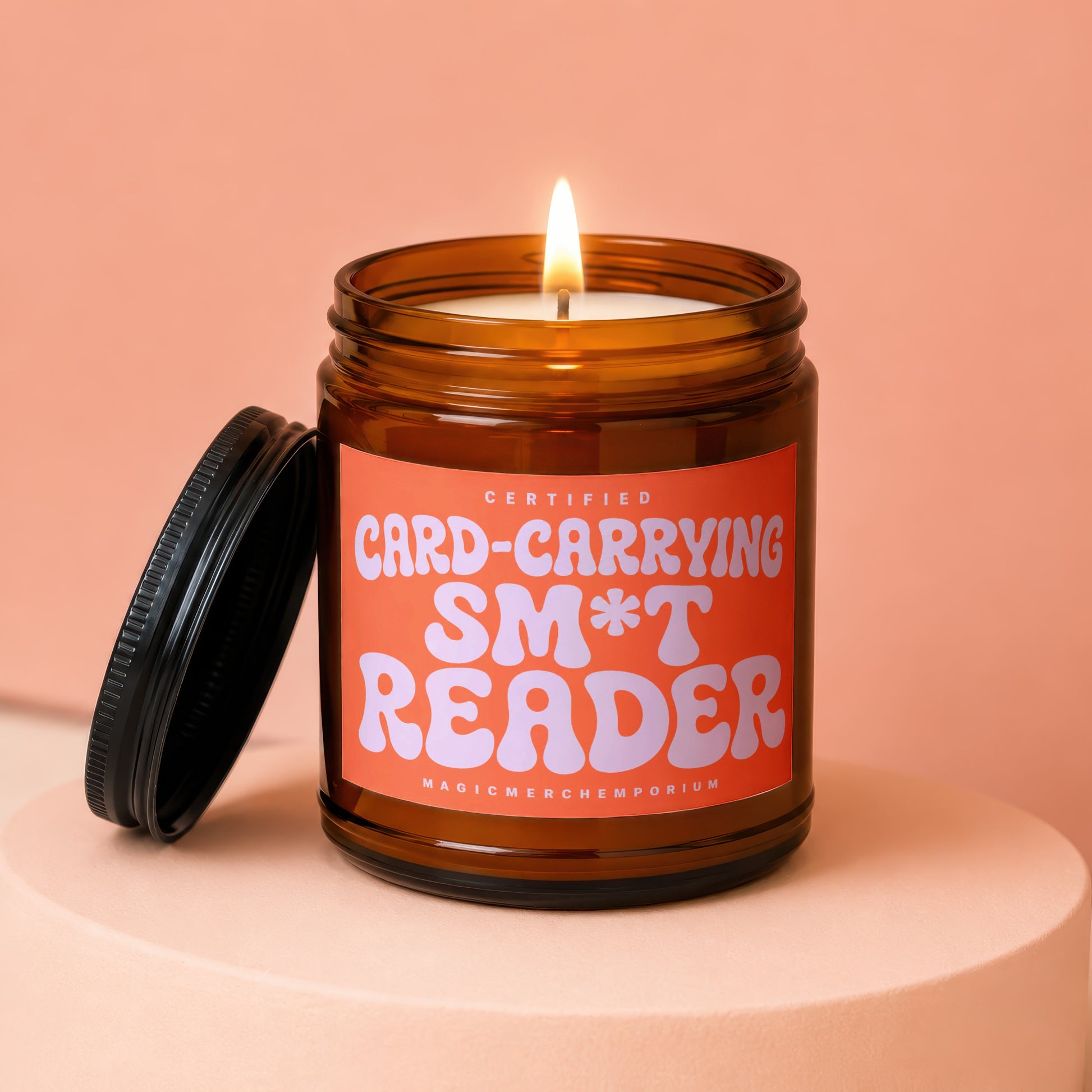Amber jar candle with orange label reading "CERTIFIED CARD-CARRYING SM*T READER".