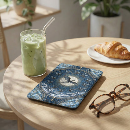 An e-reader case with a blue celestial dragon design lies on a wooden table beside a green drink, a croissant, and glasses.