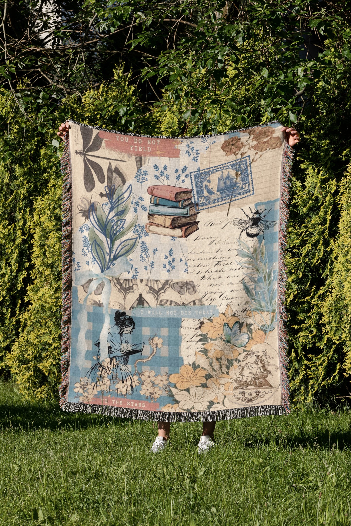 A person is holding up a colorful blanket with various illustrations and text, including a book, flowers, and a bee, in a grassy area surrounded by trees.