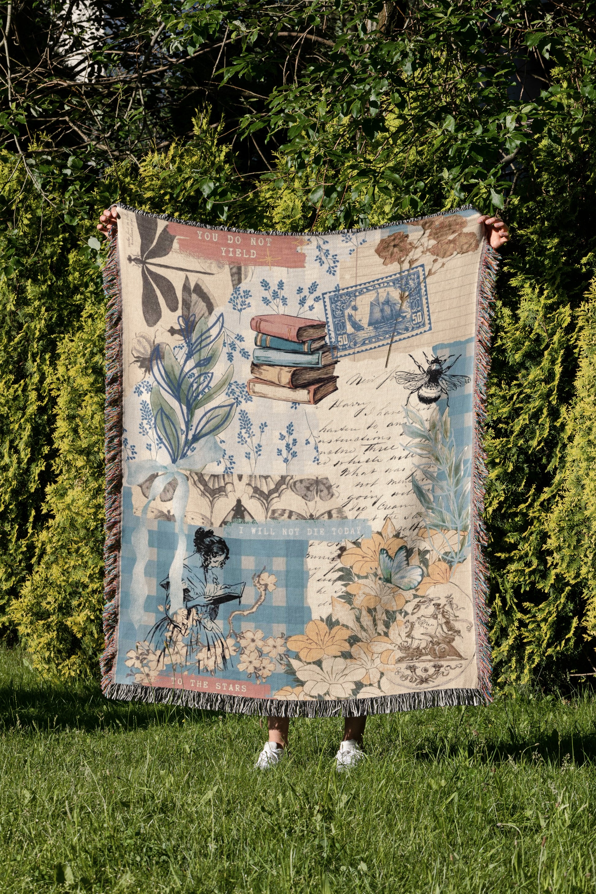 A person is holding up a colorful blanket with various illustrations and text, including a book, flowers, and a bee, in a grassy area surrounded by trees.
